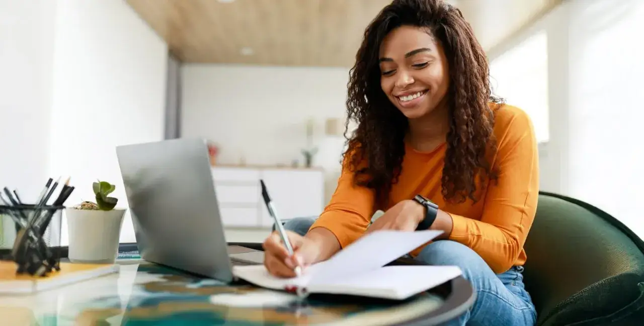 A smiling woman studies at home, researching the best online associate degree programs on her laptop and taking notes.