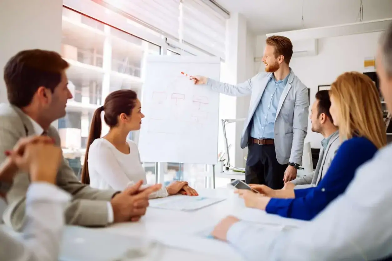 A smiling man leads a new manager training session, pointing to a flowchart on a whiteboard as attendees listen attentively.