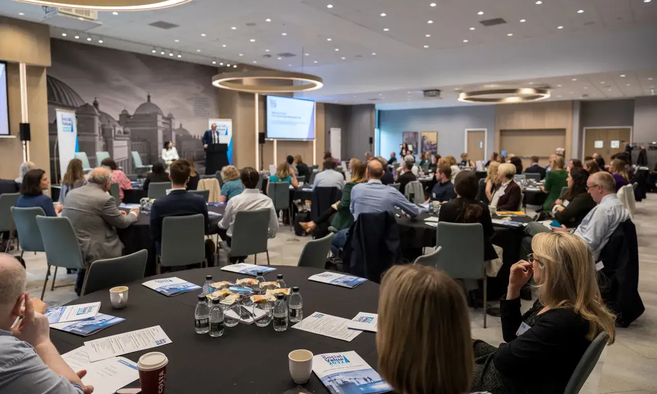 A speaker addresses an audience at a conference on philanthropic leadership. Attendees are seated at round tables, listening attentively.