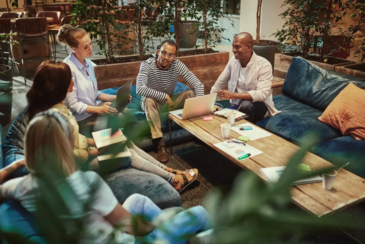 Millennials in the workplace collaborate in a relaxed, modern office setting, discussing ideas around a coffee table with laptops and papers.