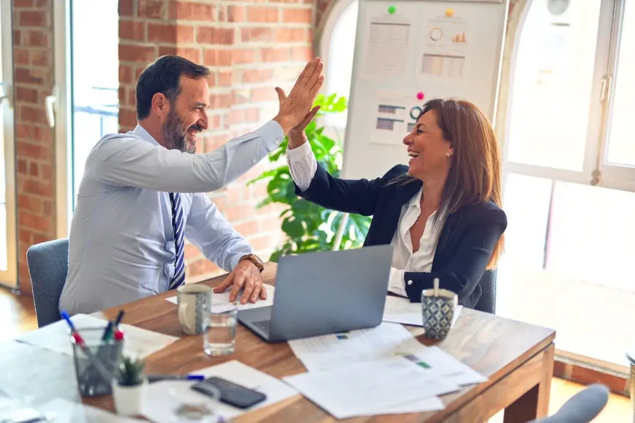 Two smiling business colleagues give each other a high five at a desk with a laptop and papers.
