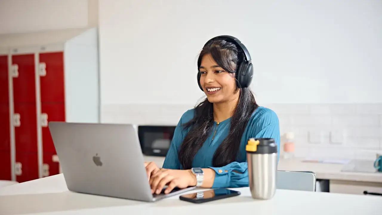 A smiling woman with headphones on is typing on her laptop, likely studying at an online tech college.