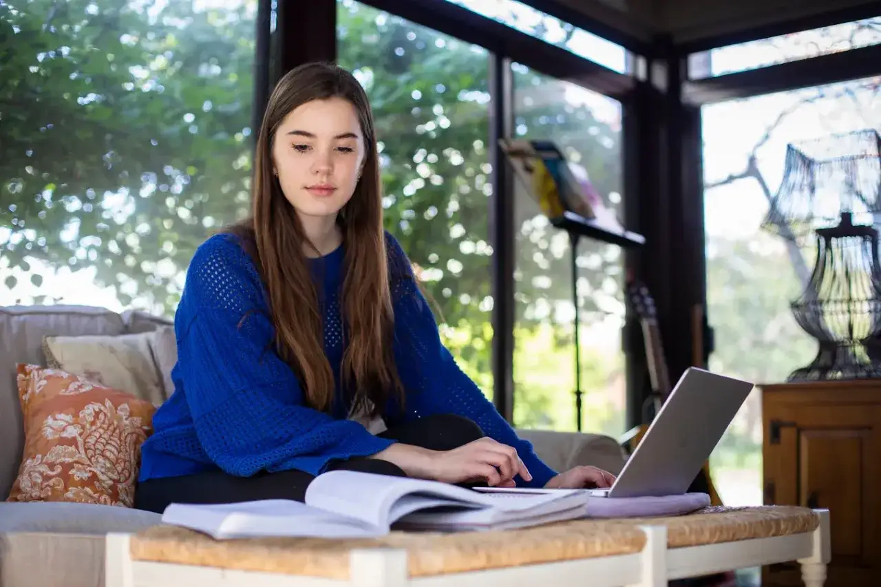 A young woman studies general education courses online, focused on her laptop while sitting on a couch with books.