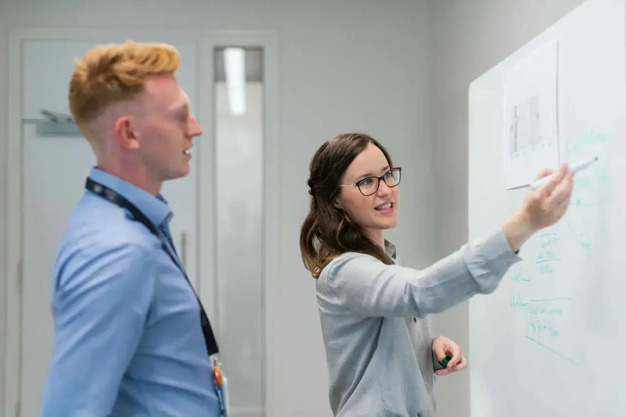 A woman with glasses leads a charter meeting, writing on a whiteboard while a man with blonde hair looks on.